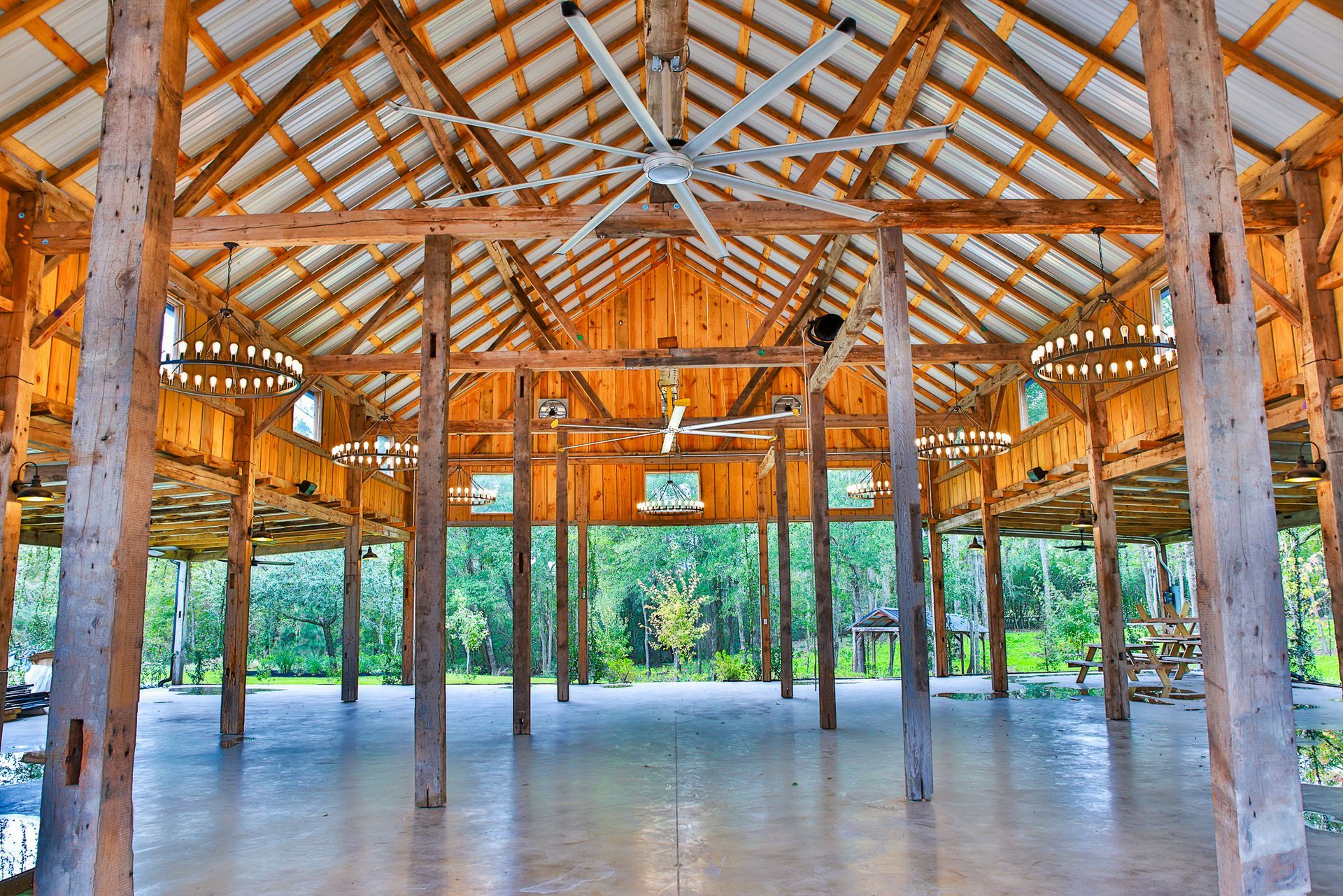 Wooden-framed open-air event space with a concrete floor, high ceilings, and surrounding trees.