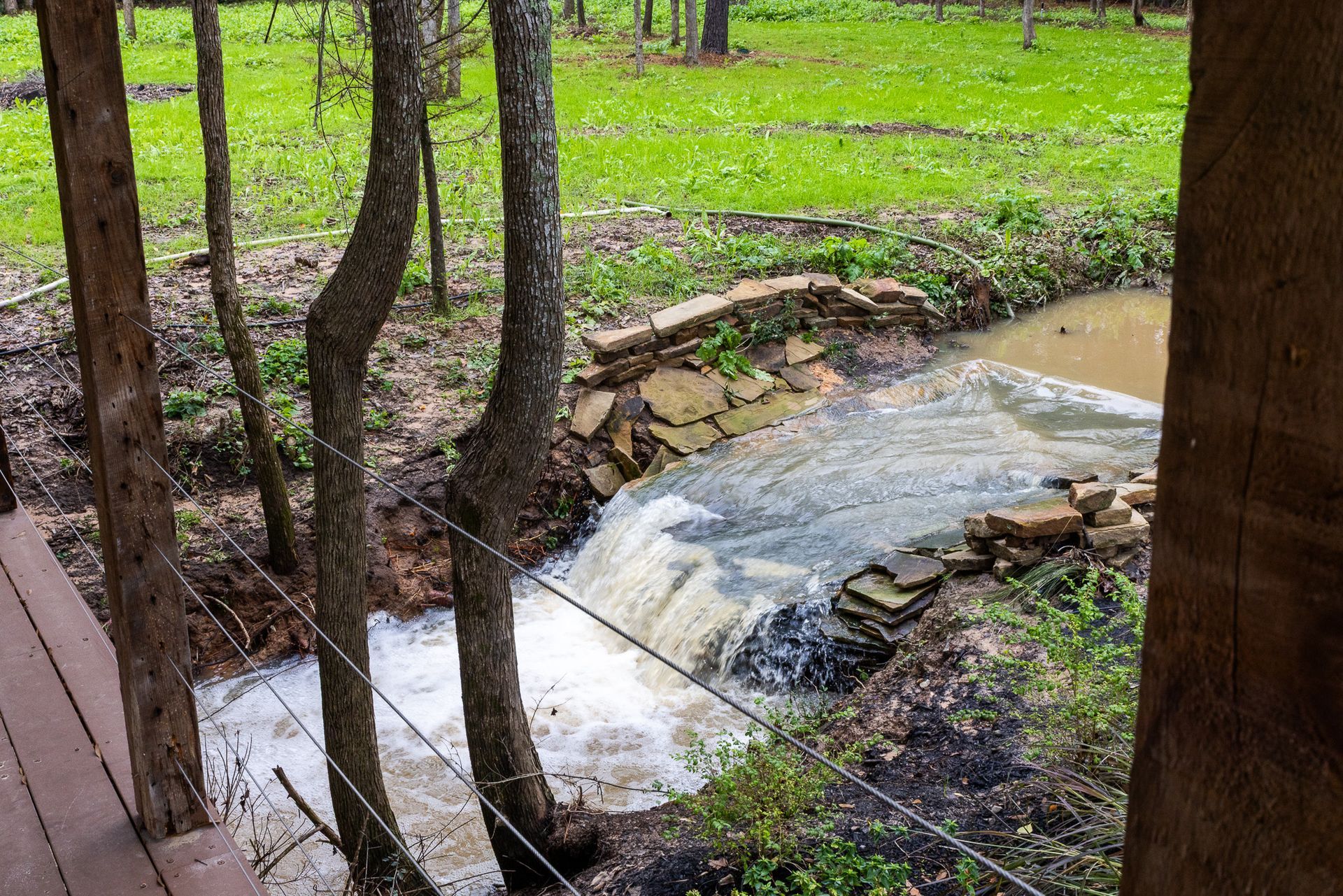 Waterfall flowing over a stone wall in a wooded area, viewed from a wooden structure.