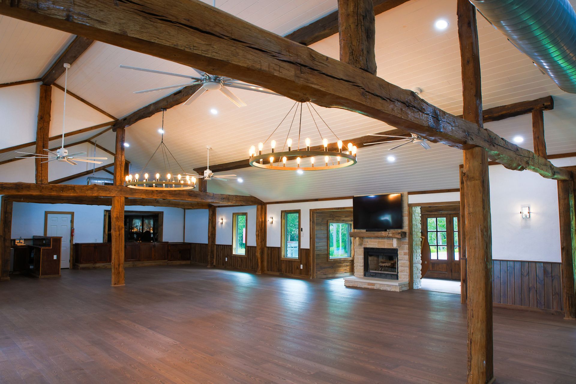 Event space with wooden beams, hardwood floor, chandeliers, fireplace, and windows.