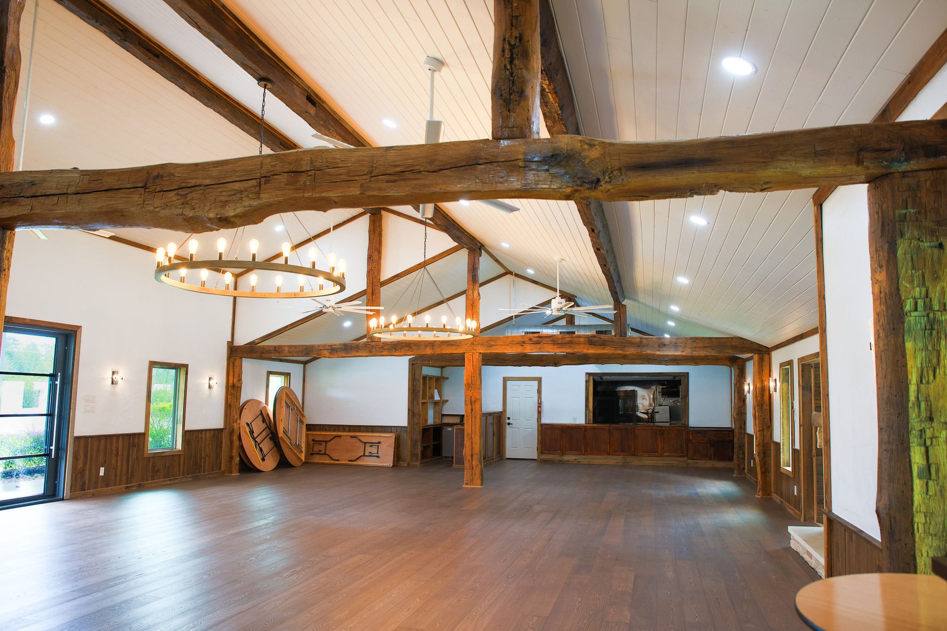 Empty event space with wooden beams, chandeliers, and wood flooring.