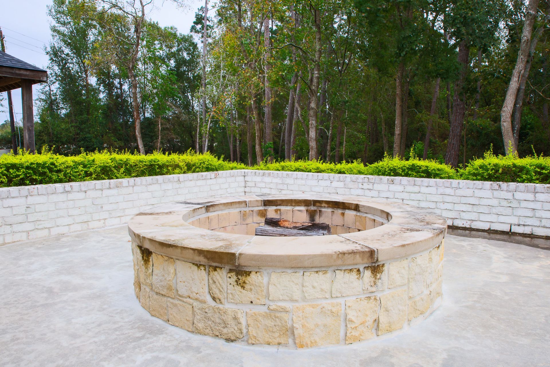 Stone fire pit on a concrete patio, surrounded by white brick walls and trees.