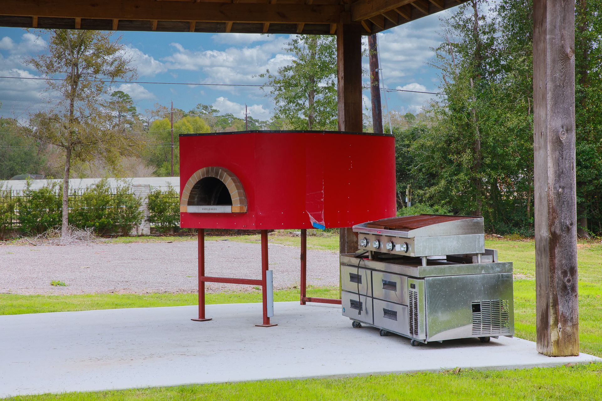 Red outdoor pizza oven and stainless steel grill under a wooden shelter on a concrete slab.