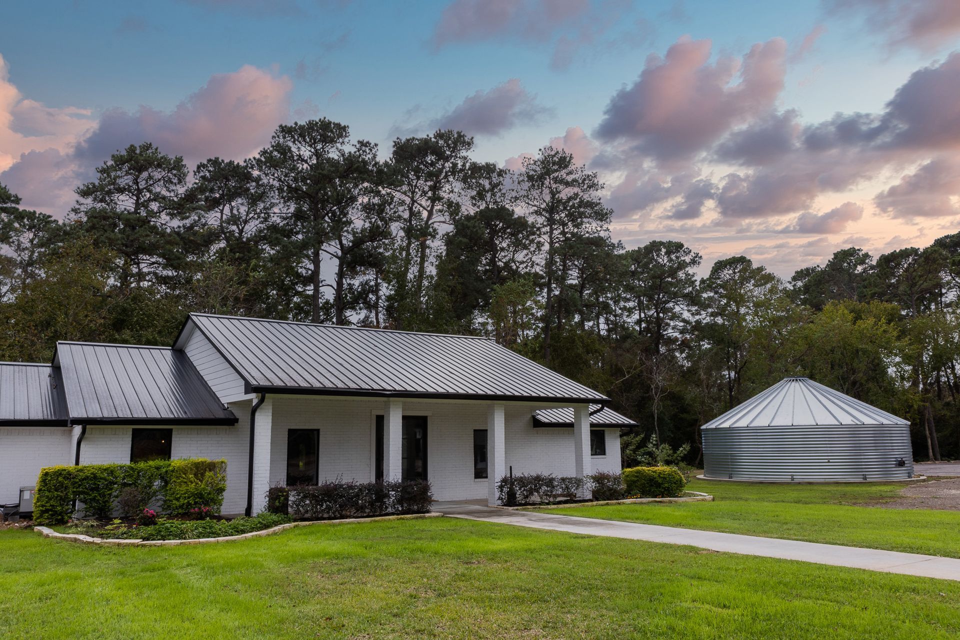 White house with metal roof and silo on a green lawn with a tree-lined backdrop under a sunset sky.