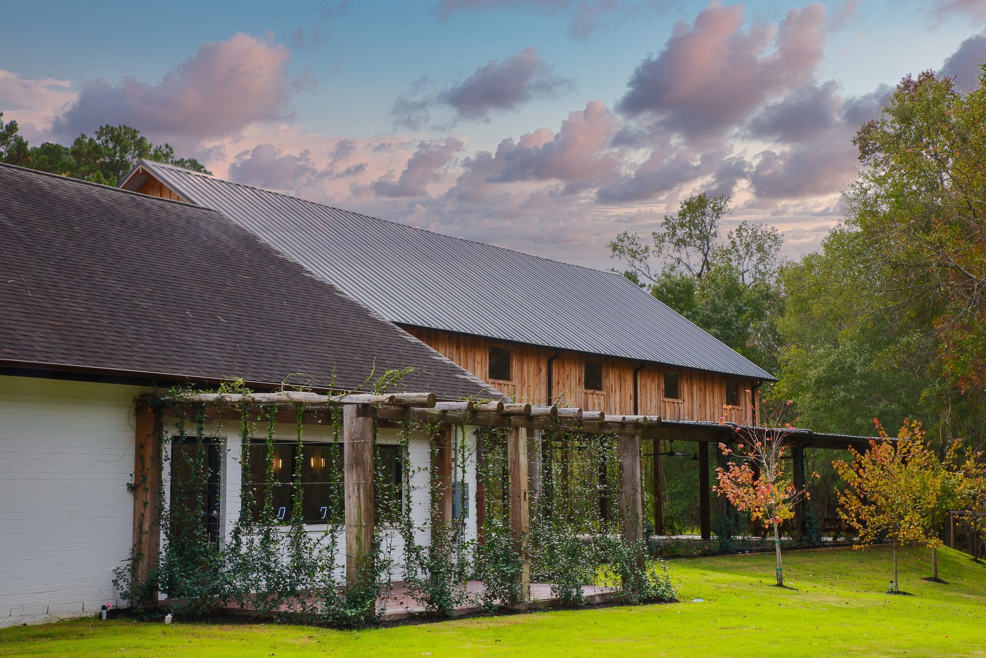 Wooden barn and white building with a covered porch; green lawn and trees under a cloudy sky.