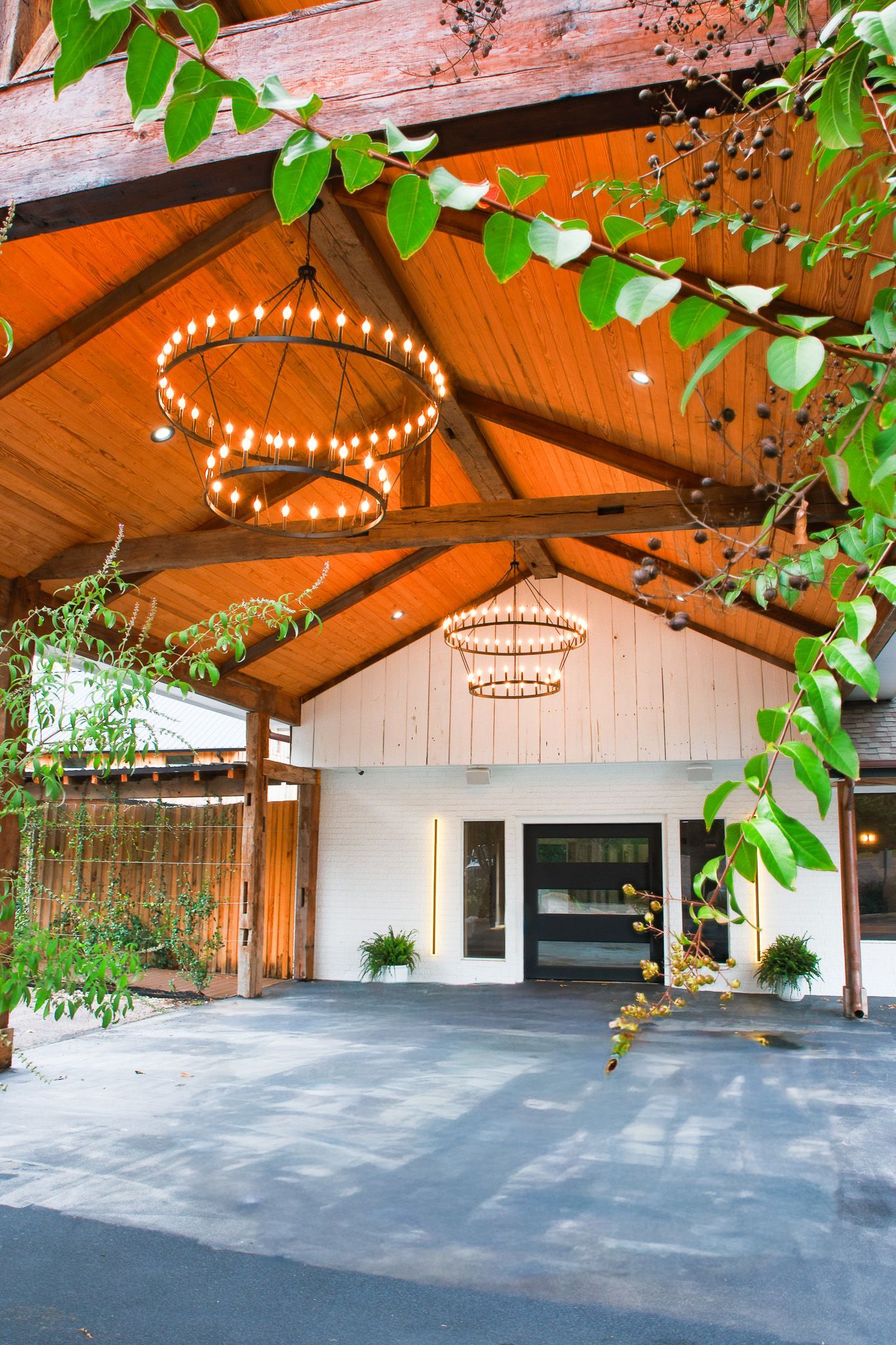 Exterior view of a building with a wooden pergola, lights, and a white facade. Green foliage frames the image.