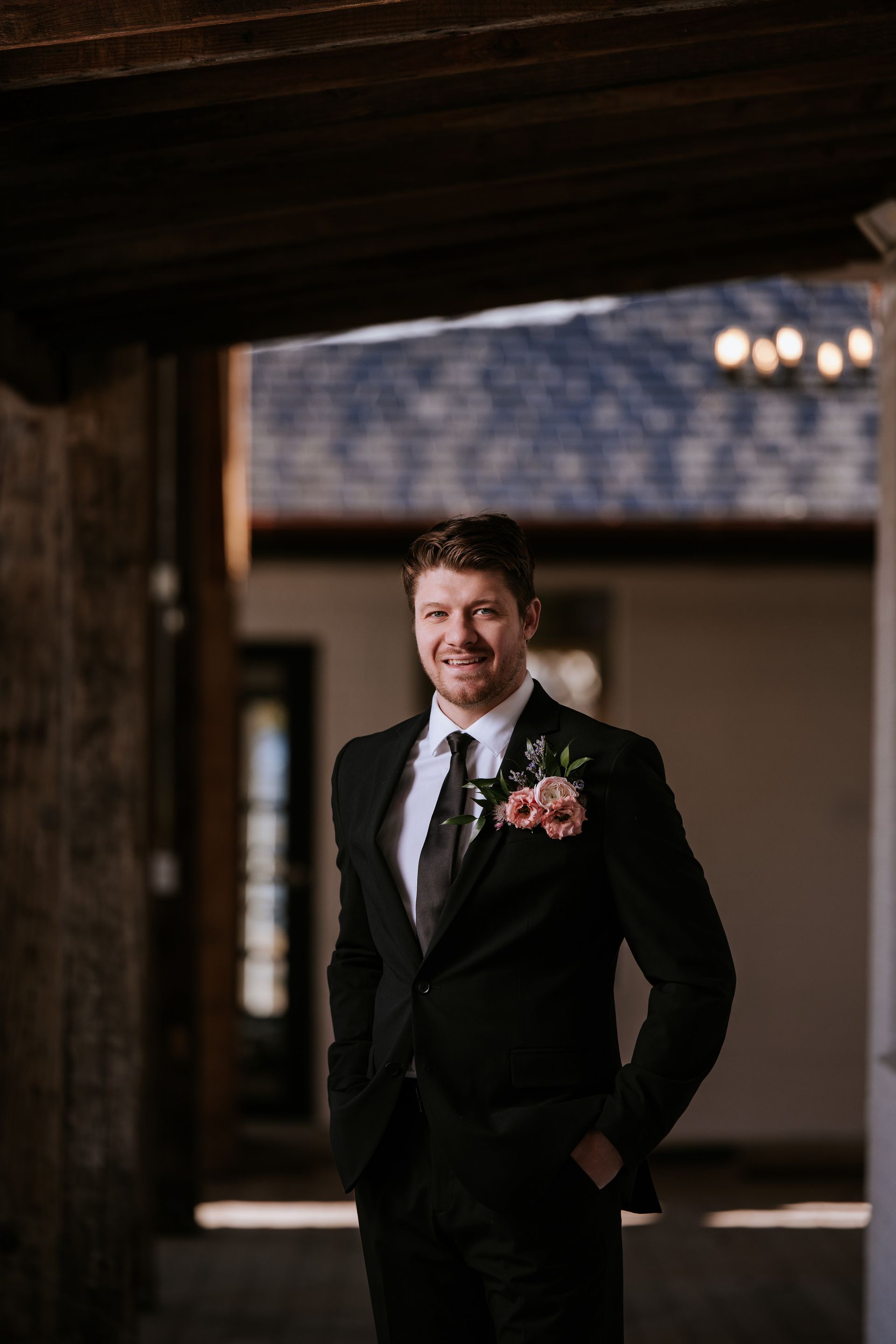 Man in black suit smiles, standing outdoors in a covered area, floral boutonniere.