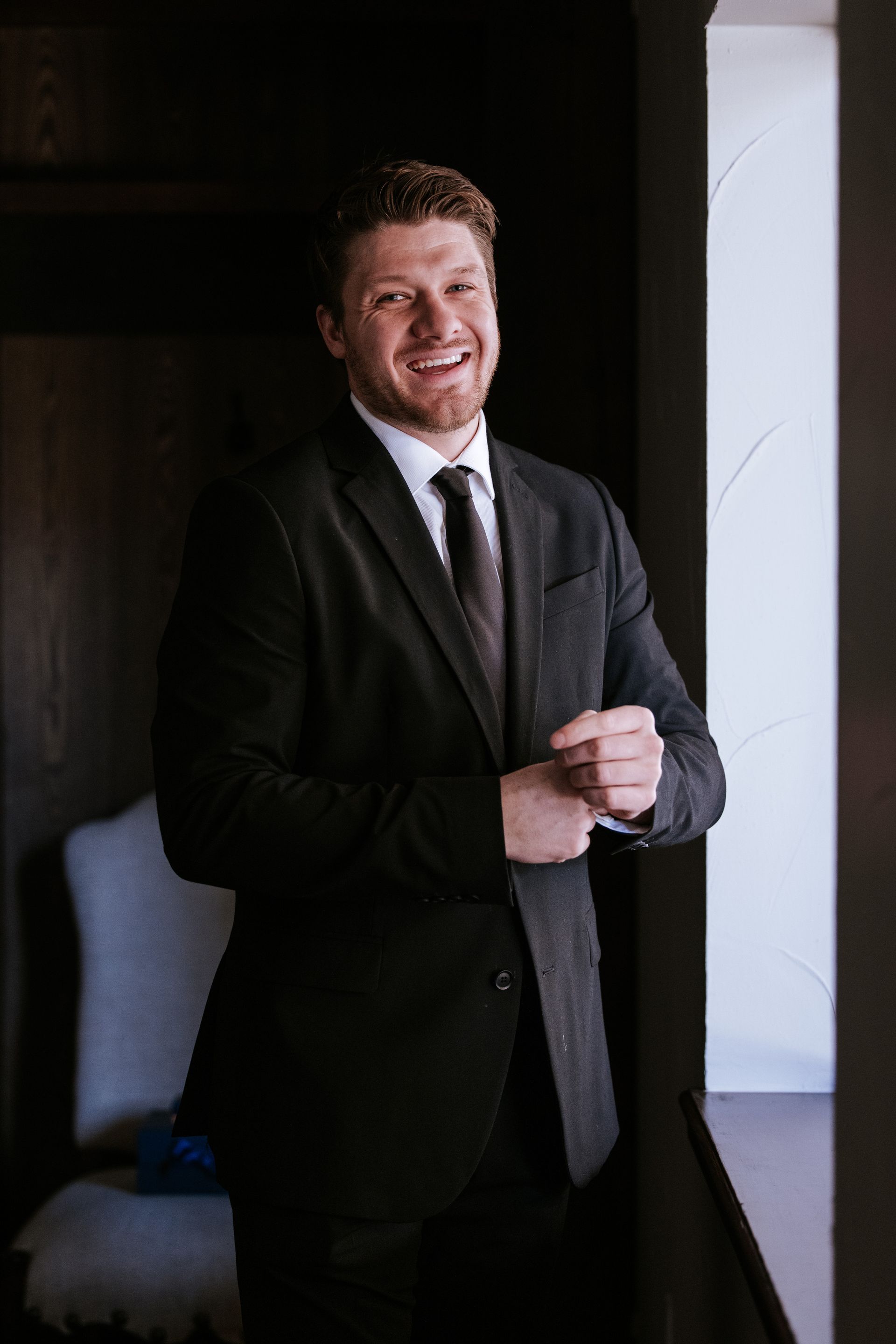 Man in a dark suit smiles, adjusting his cuff near a doorway, with a chair visible.