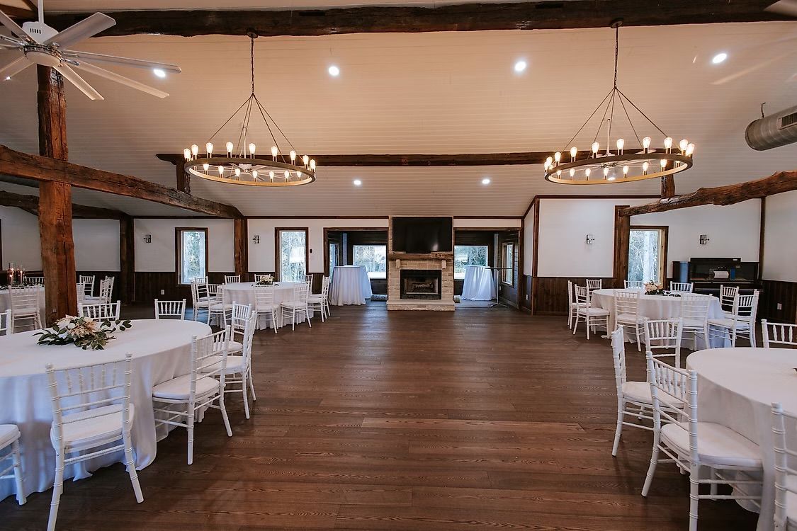Ivey Cabin with round tables set for a wedding; wooden floor, chandeliers, white chairs, fireplace.