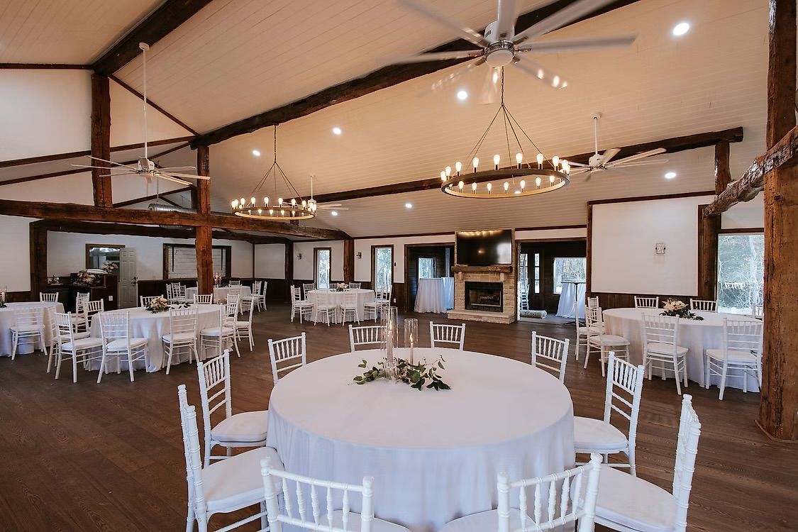 Ivey Cabin venue with white round tables set for a wedding; white chairs, wooden beams, chandeliers, and fireplace.