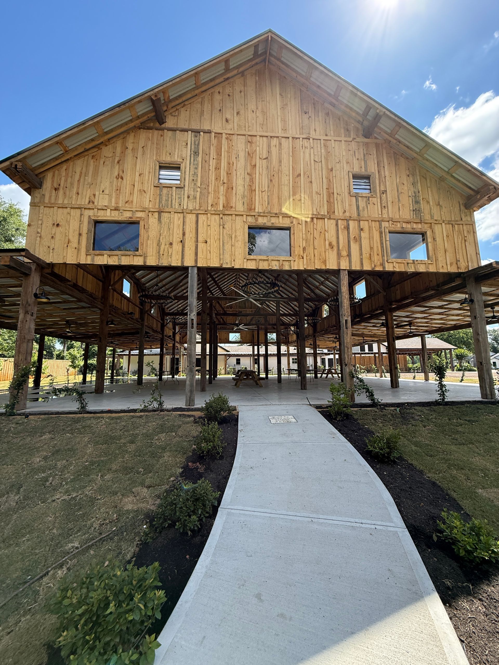 Wooden two-story structure on stilts with a pathway leading to the entrance; outdoor setting with greenery and blue sky.