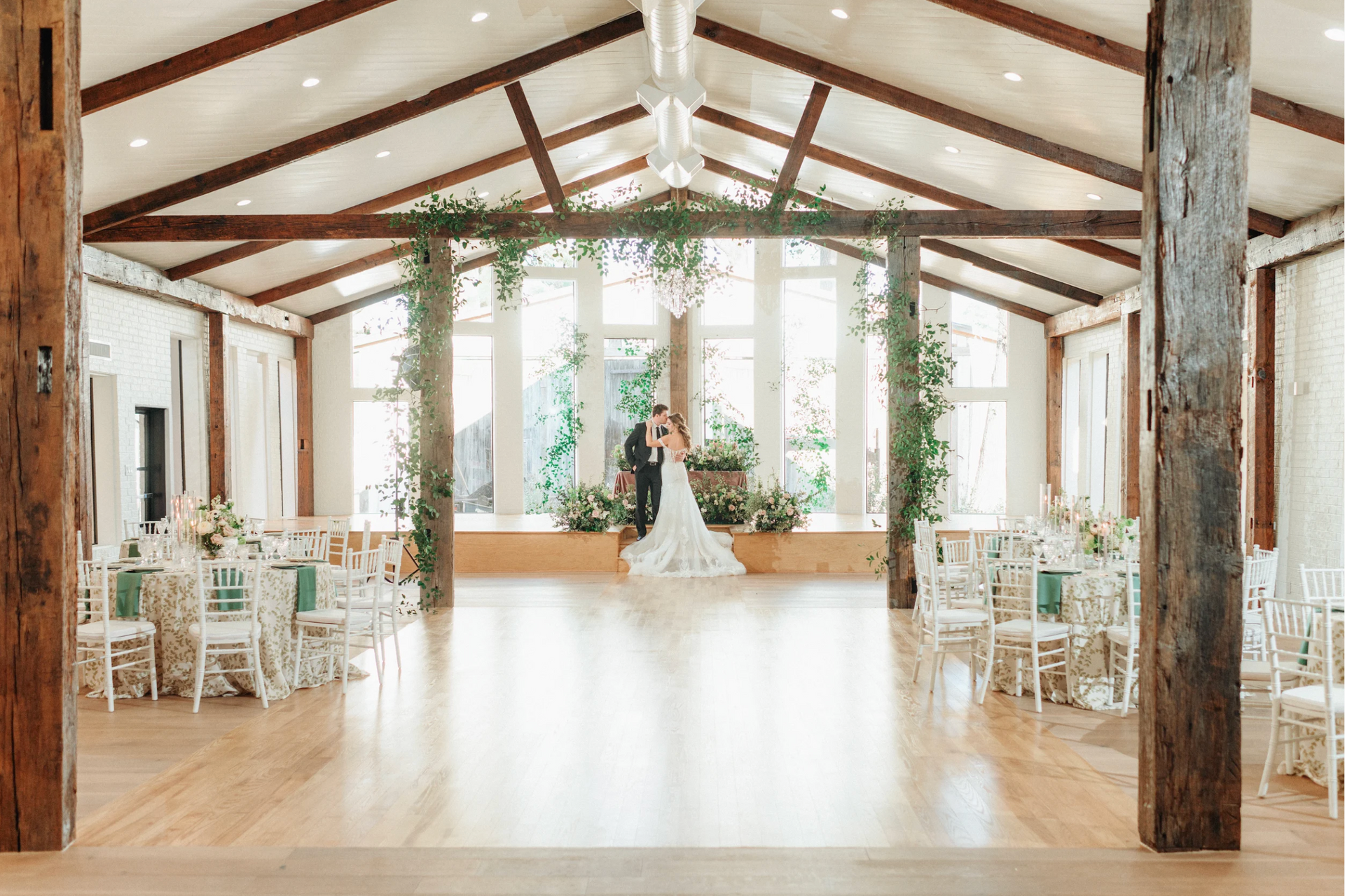 Wedding reception hall with bride and groom, wooden beams, floral arch, and white chairs.