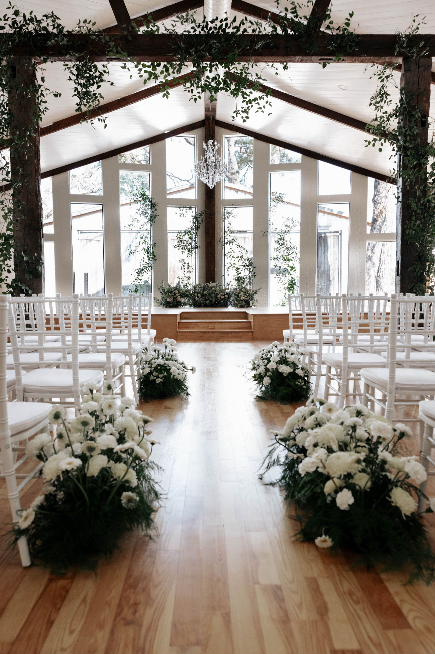 Wedding ceremony setup with white chairs, floral arrangements, and greenery in an indoor venue with large windows.