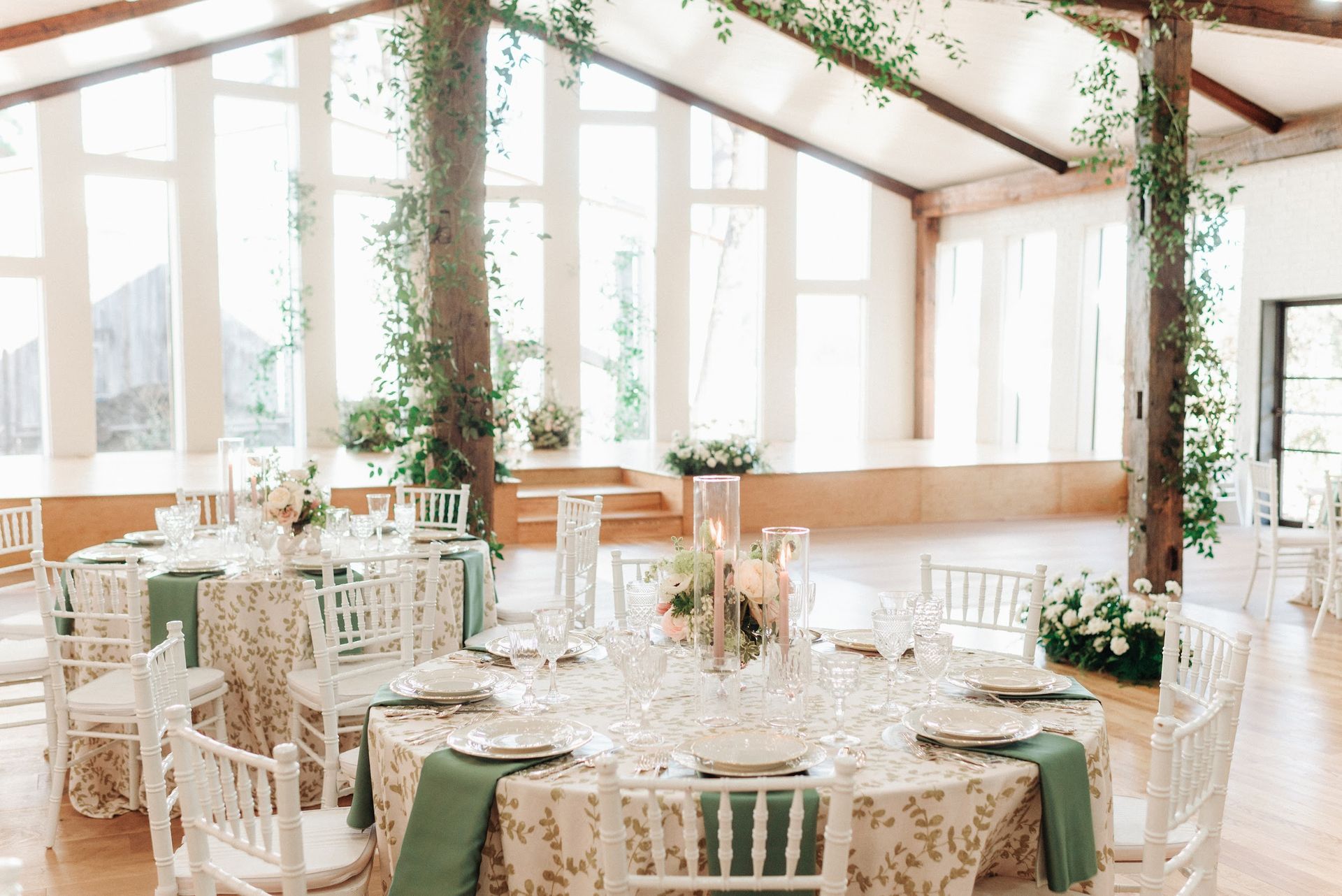 Elegant banquet hall set for a wedding; round tables with floral linens and green napkins, tall windows.