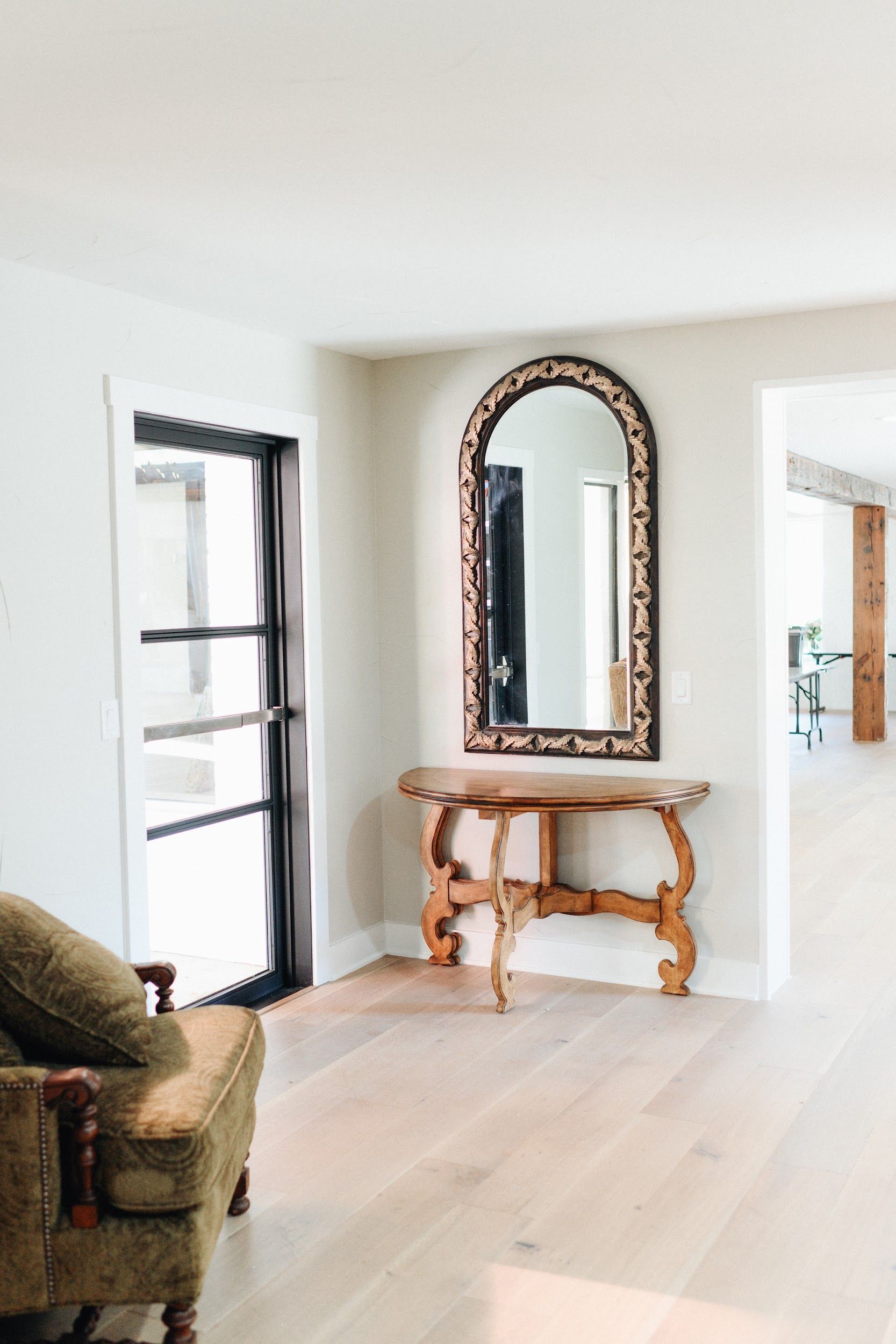 Entryway with arched mirror above wooden console, black-framed door, and light wood flooring.