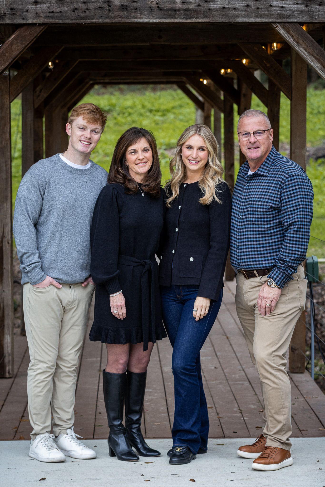 Family of four posing under wooden structure. Two women, two men, smiling. Outdoors, neutral background.