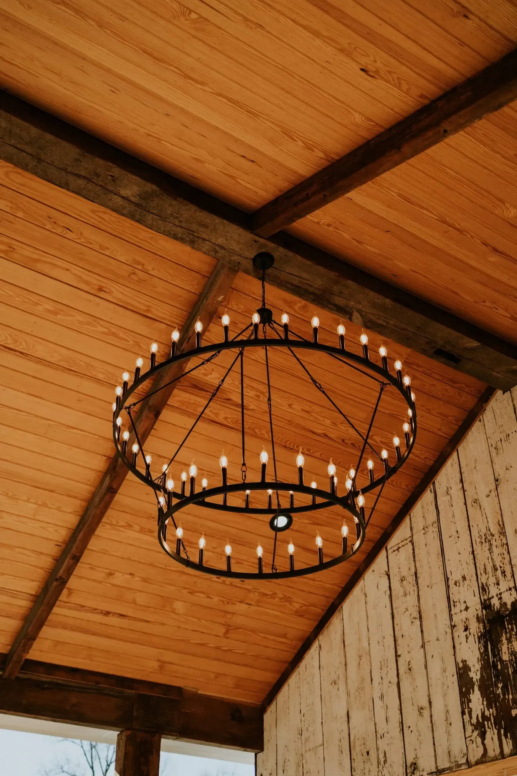Two-tiered black chandelier hanging from a wood plank ceiling with brown beams.