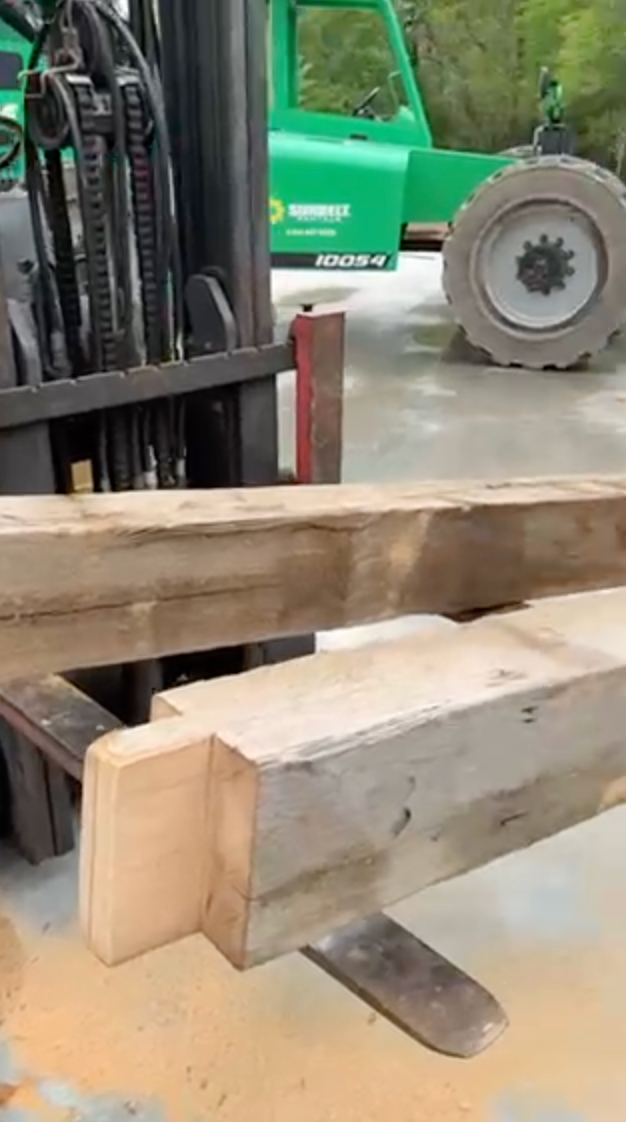 Forklift lifting a large wooden beam, green vehicle in the blurred background.