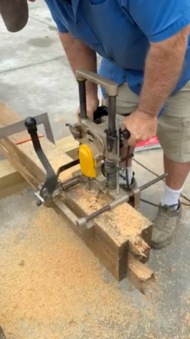 Person using a mortise machine on a wooden beam, creating a joint. Outdoors, with wood shavings visible.