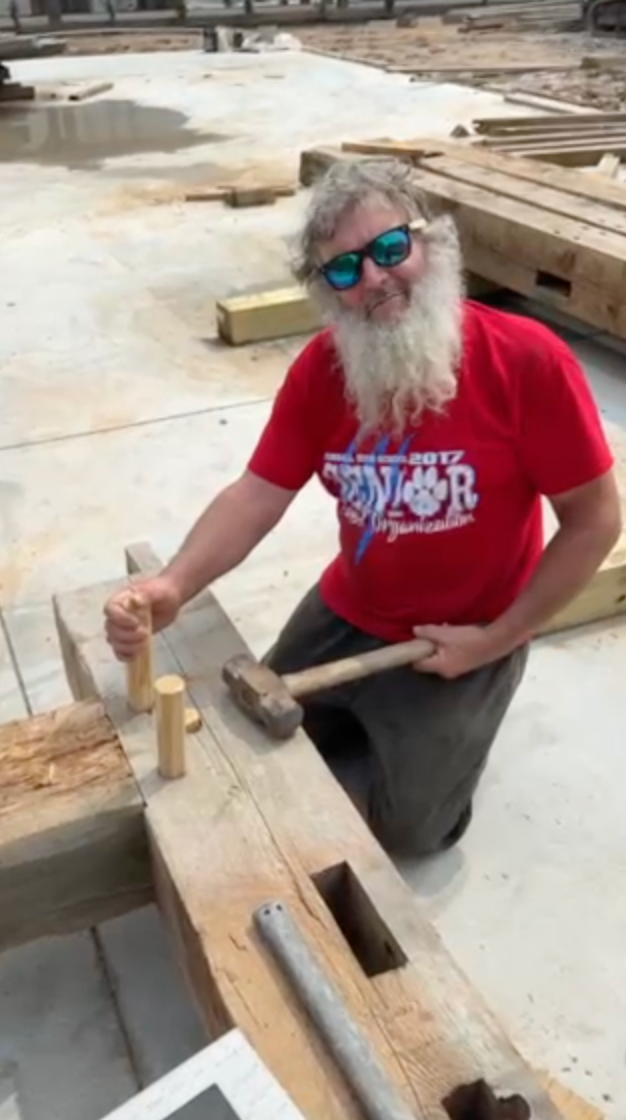 Man in red shirt and sunglasses hammering pegs into wooden beams at a construction site.
