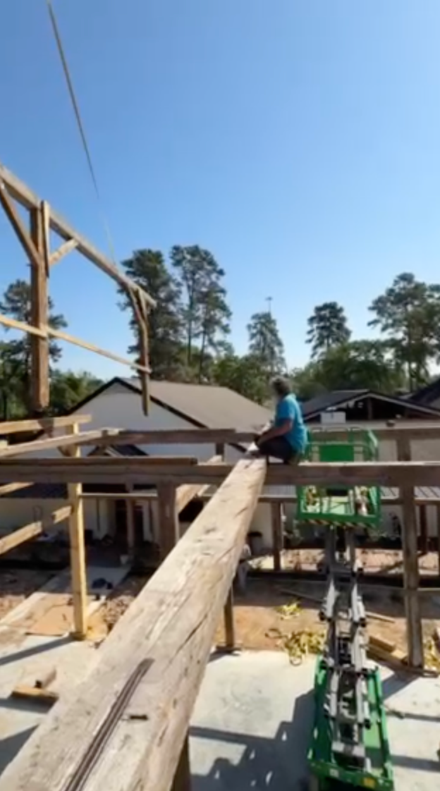 Construction worker seated on a wooden beam, working on a frame. Bright blue sky overhead.