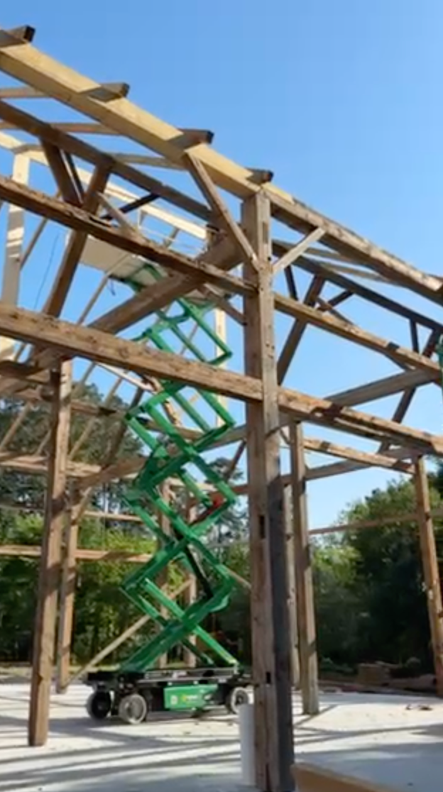 Wooden barn frame under construction with a green lift, against a blue sky.