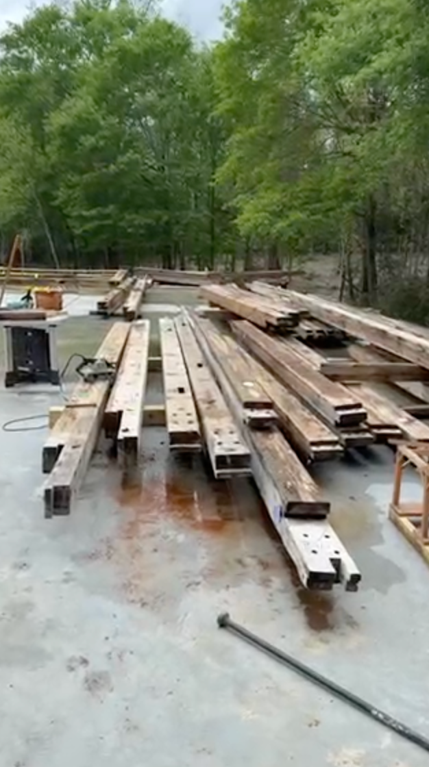 Wooden beams on a concrete surface, with trees in the background, a saw and building materials.
