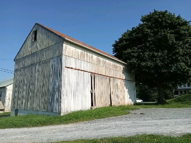 Old, weathered white barn with large sliding doors, set on a gravel path near a green tree under a blue sky.