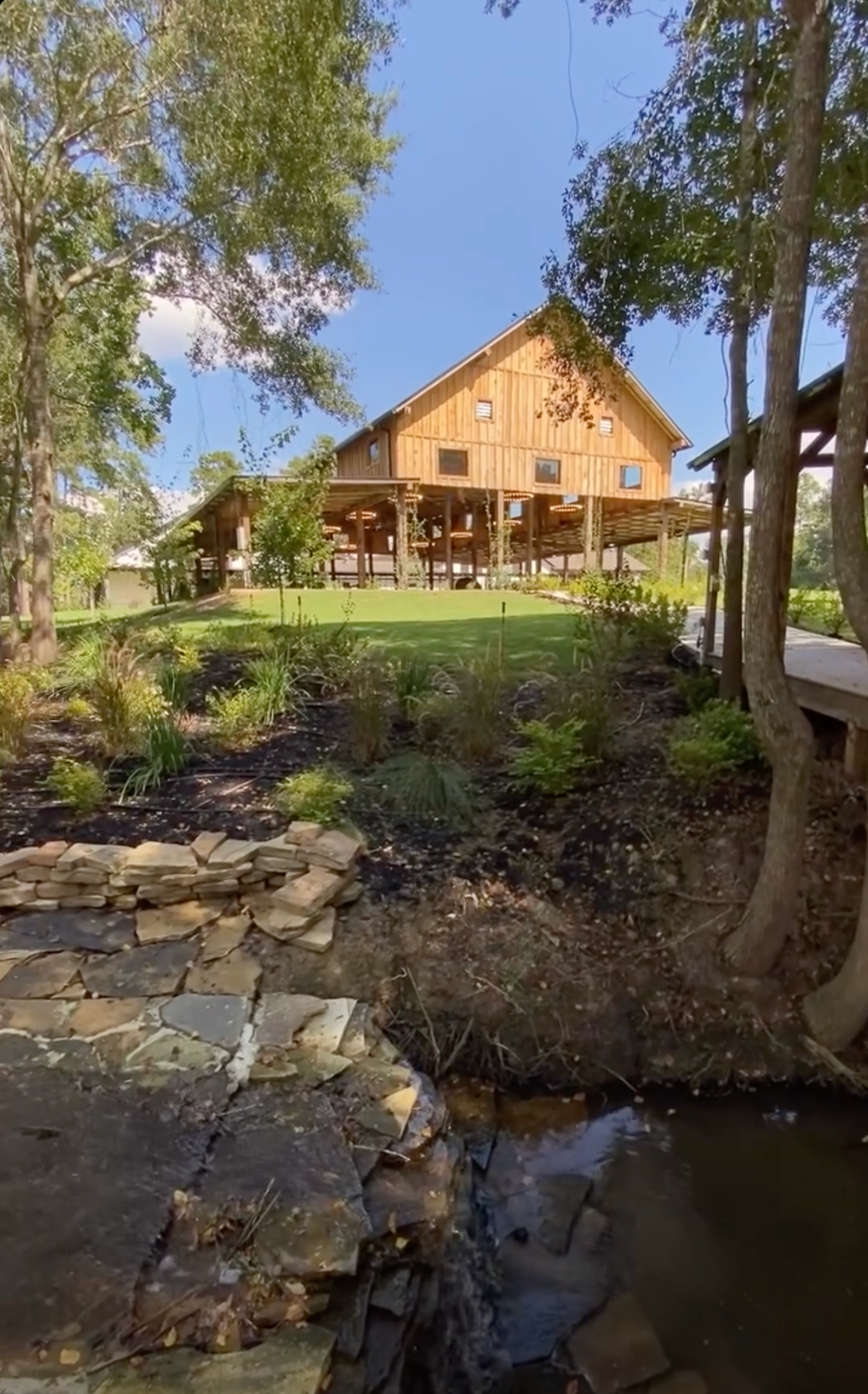 Wooden barn with porch, near stream, set in a natural landscape with trees and greenery.