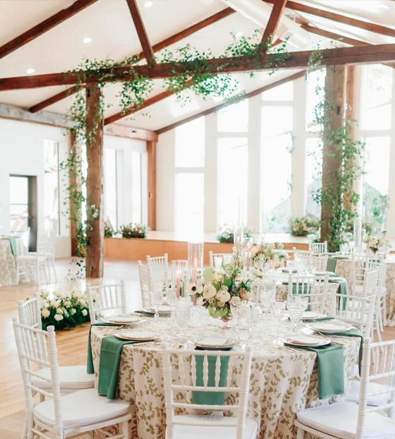 Wedding reception: Round tables set with floral linens, green napkins, white chairs in a bright venue with tall windows and wood beams with greenery.