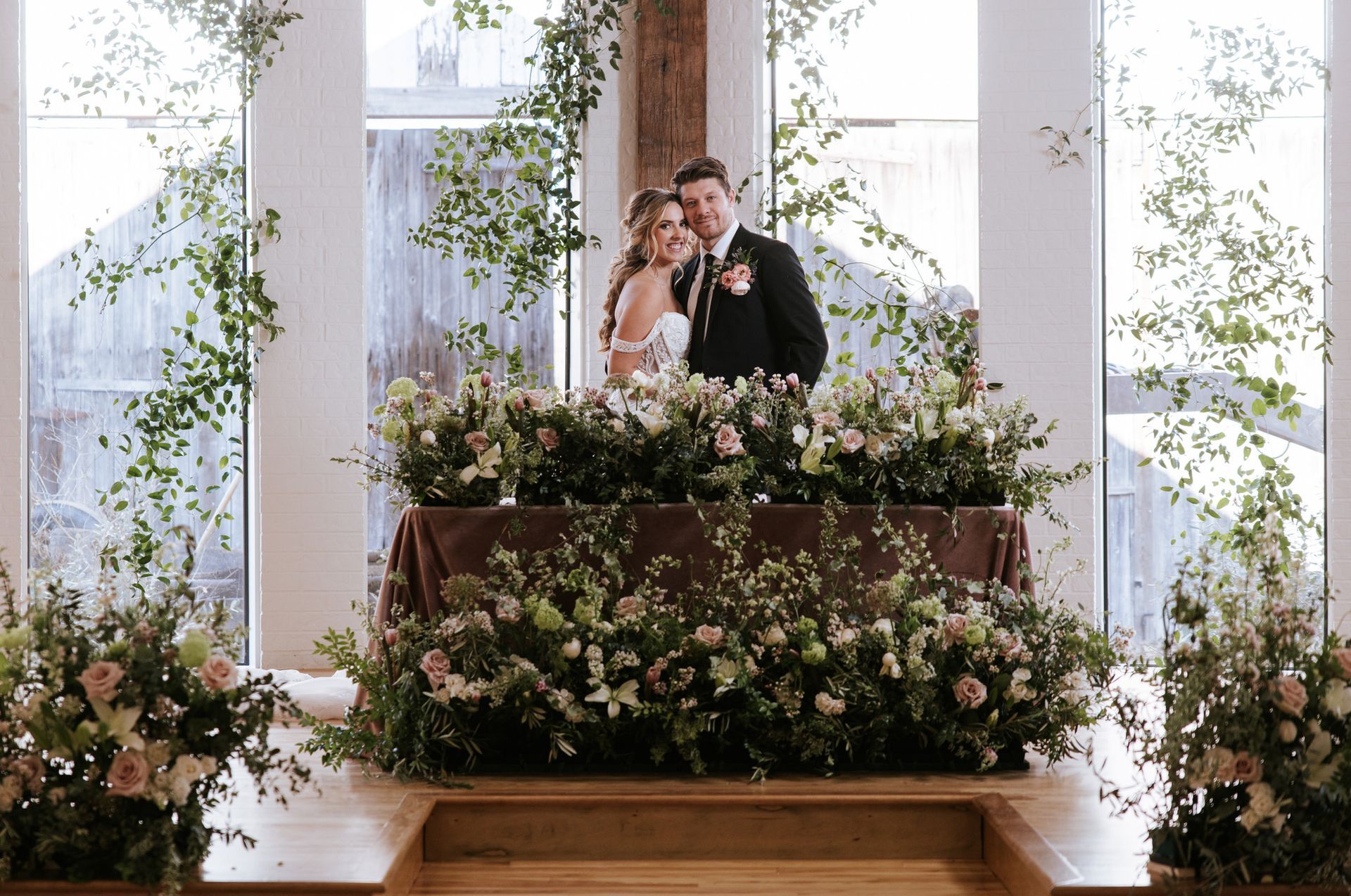 Couple standing behind a flower-covered table, inside a building with windows, adorned with greenery.