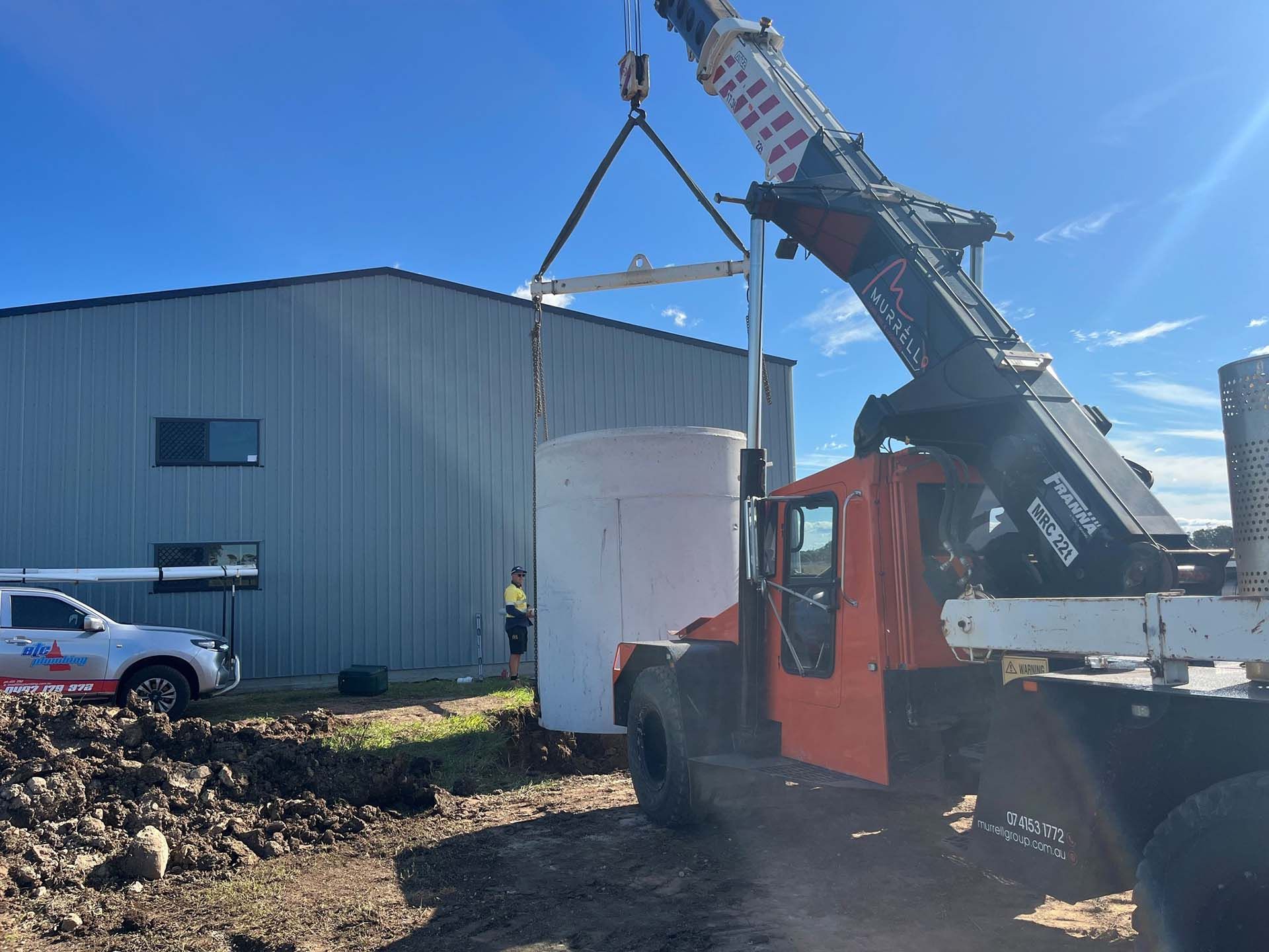 A crane lifts a large white tank near a gray commercial building