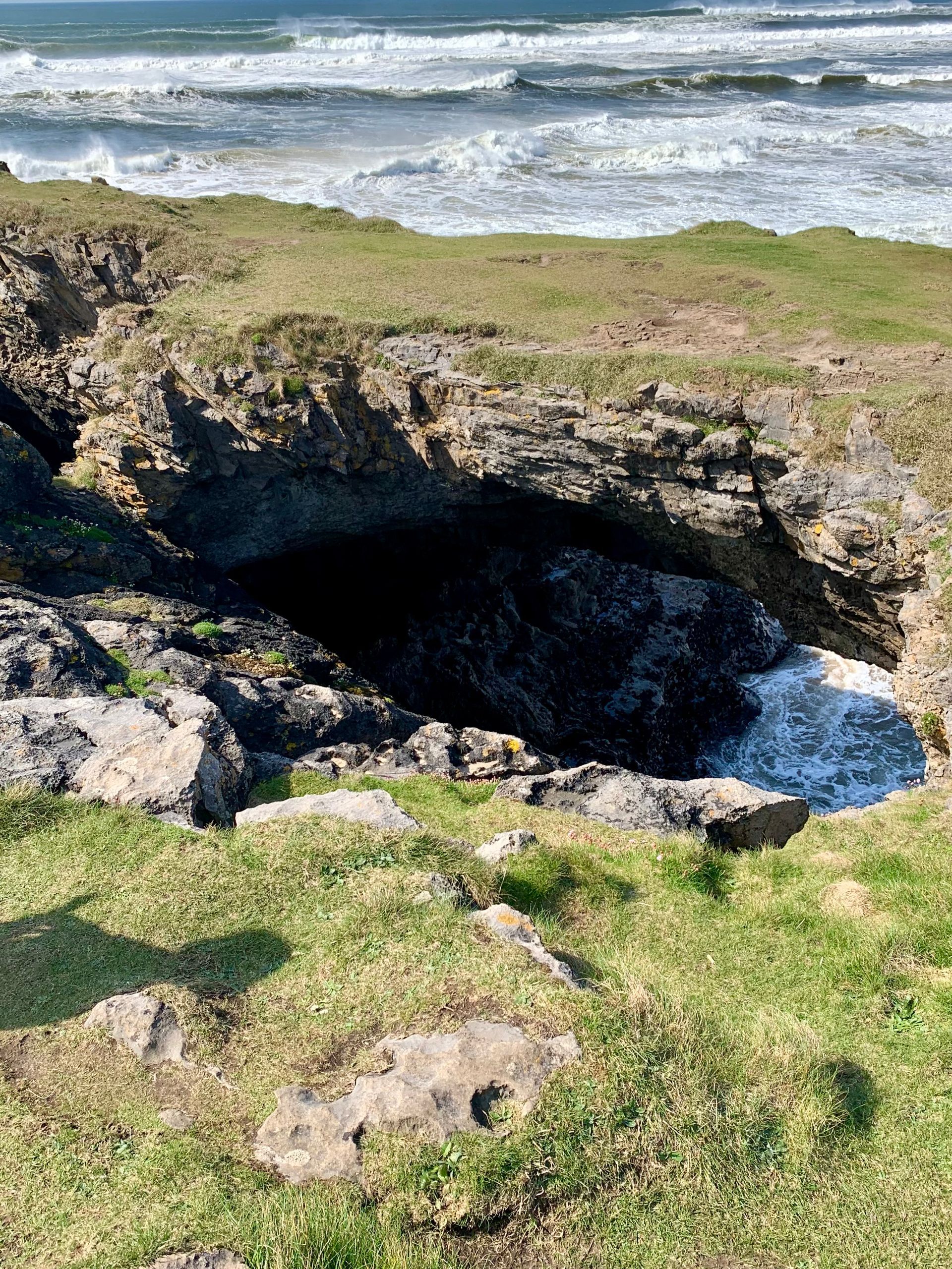Rocky coastline with waves crashing into a dark cave entrance. Green grass and ocean in the background. Fairy Bridge, Bundoran, Donegal, Ireland