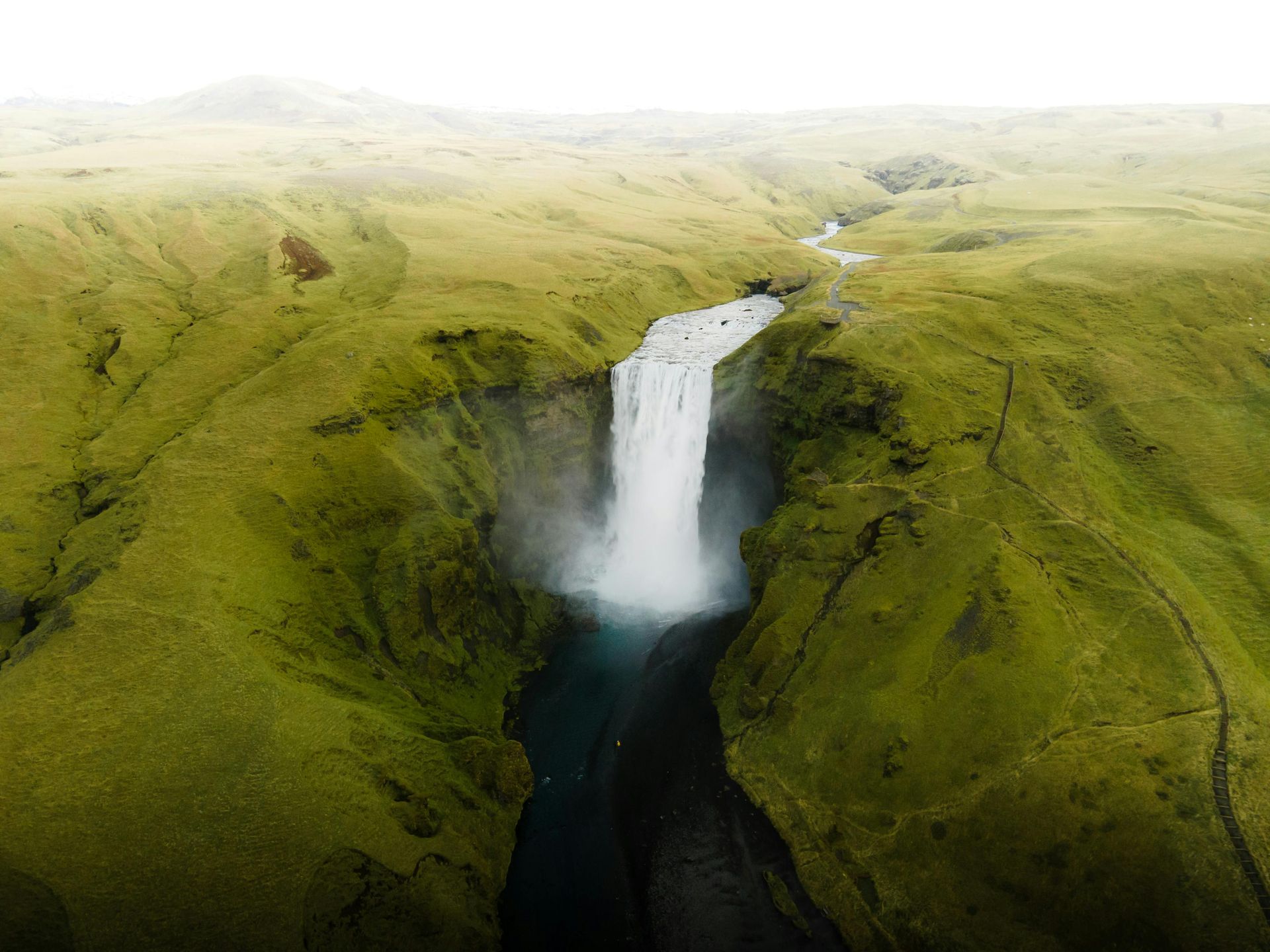 An aerial view of Skógafoss waterfall in Iceland cascading into a river through a lush, green Icelandic landscape.