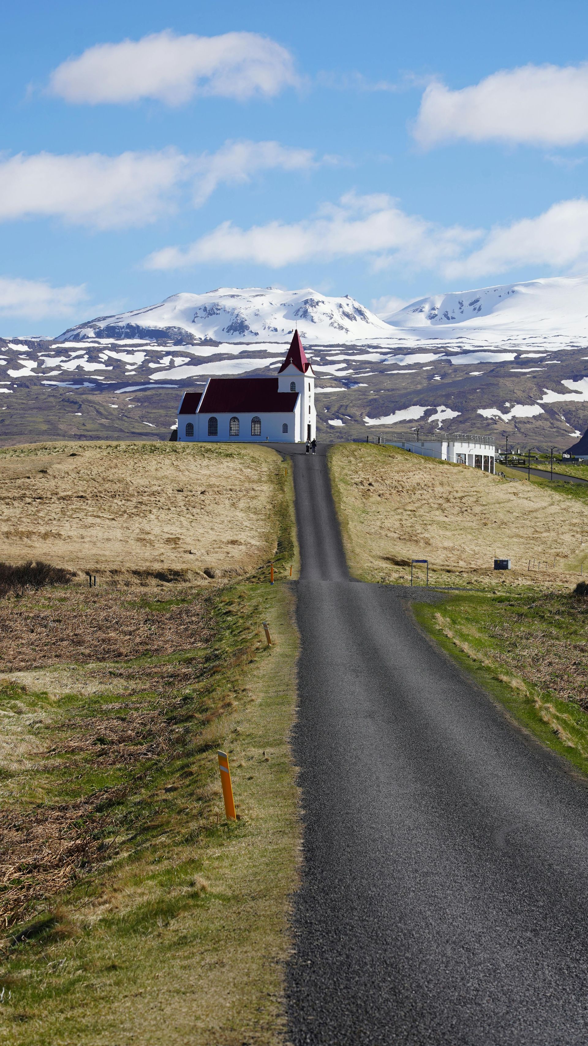 A long gravel road leads to a white church with a red roof, set against a backdrop of snowy mountains under a blue sky in Iceland
