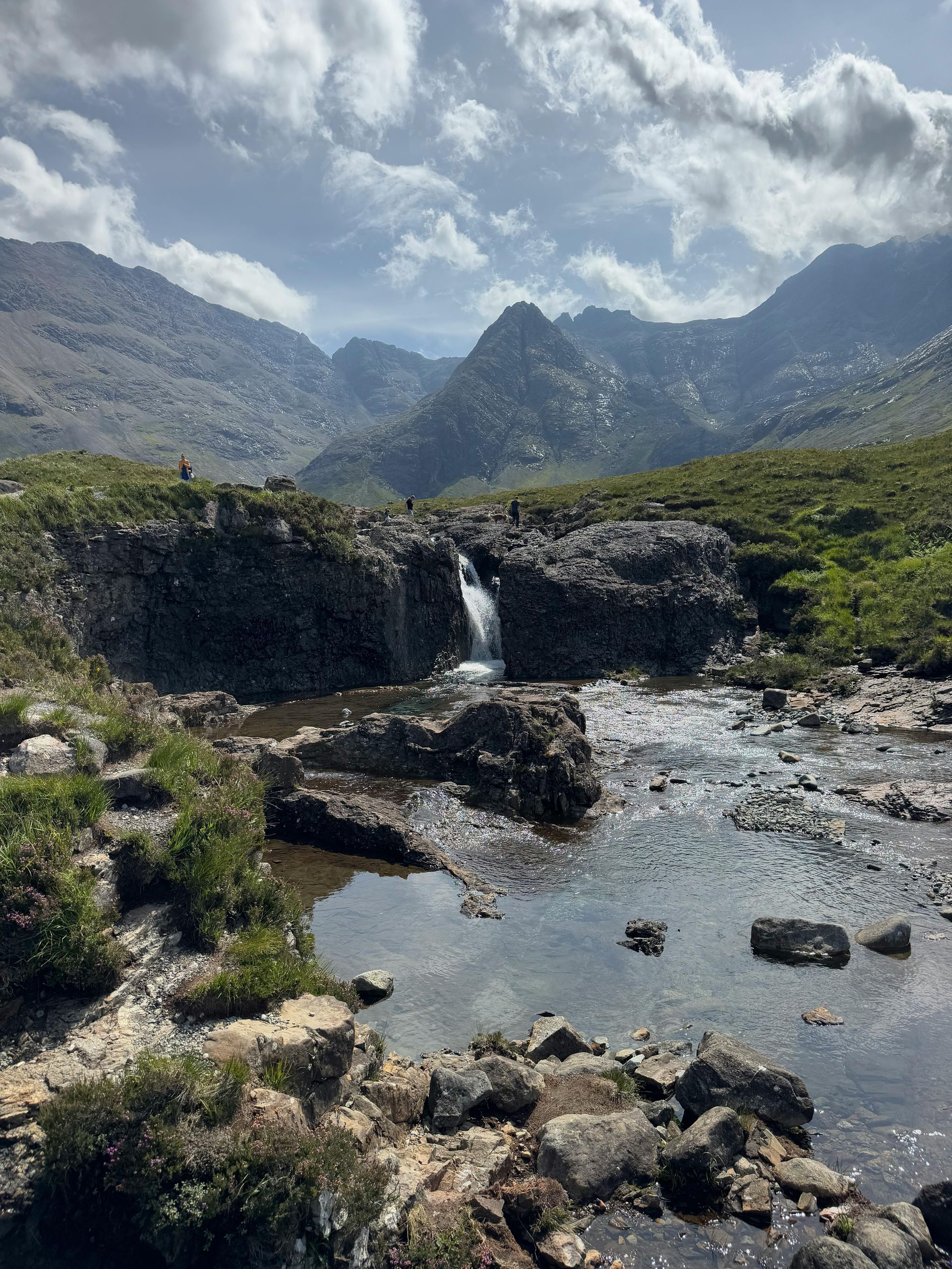 Waterfall cascades into a rocky river. Mountains rise in the background under a cloudy sky.
