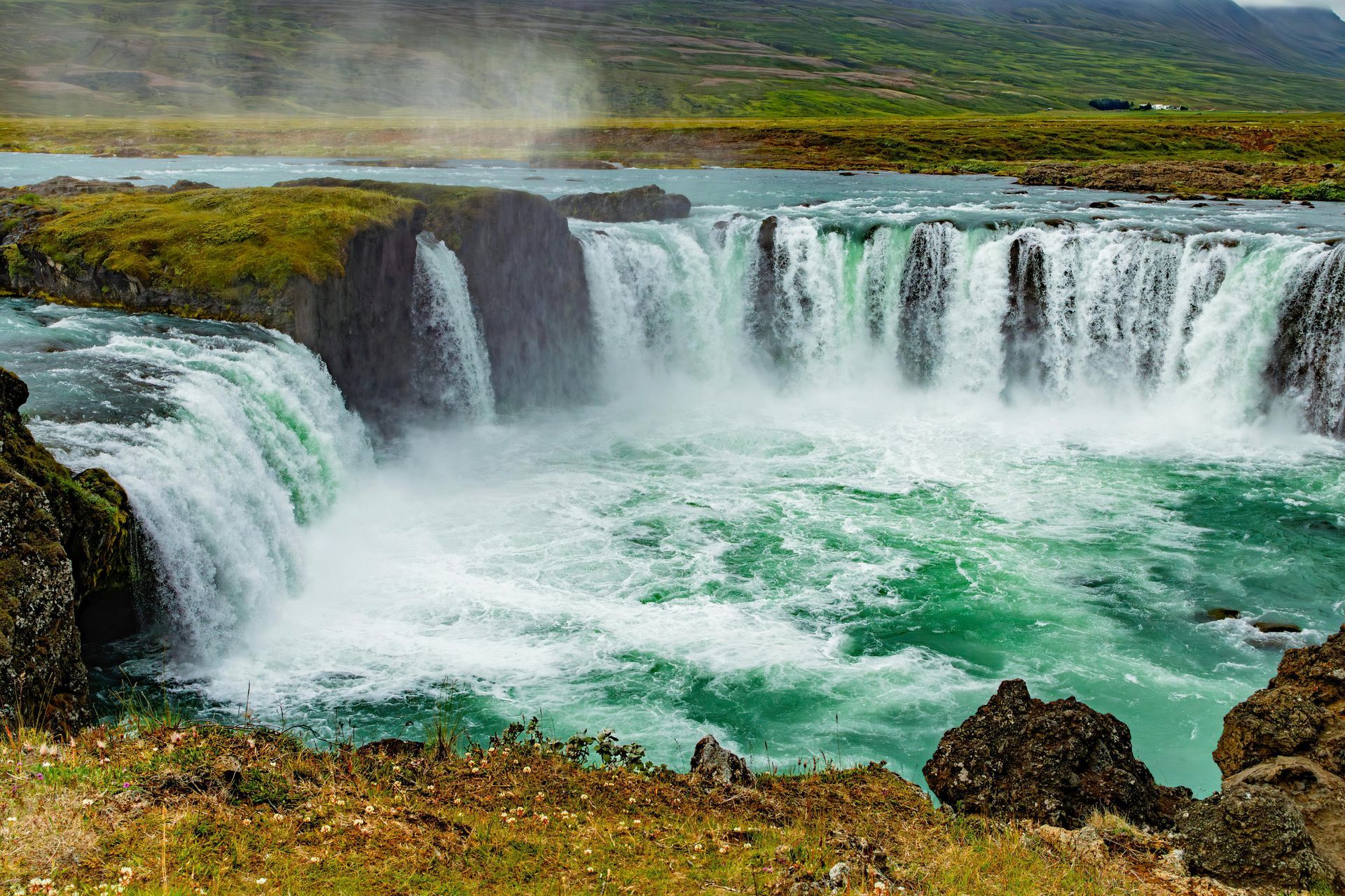 A wide, tiered waterfall in Iceland with turquoise water cascading over dark, mossy rock ledges into a pool below.