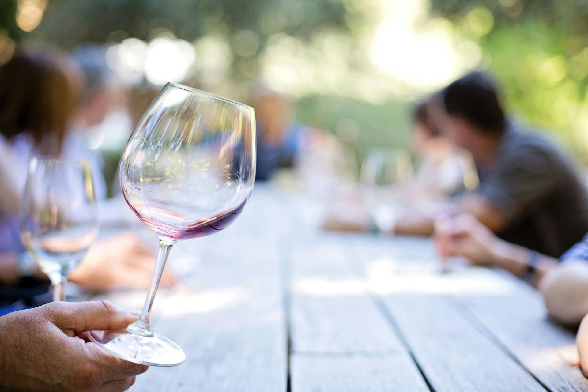 Person holding wine glass, blurred outdoor table with people.