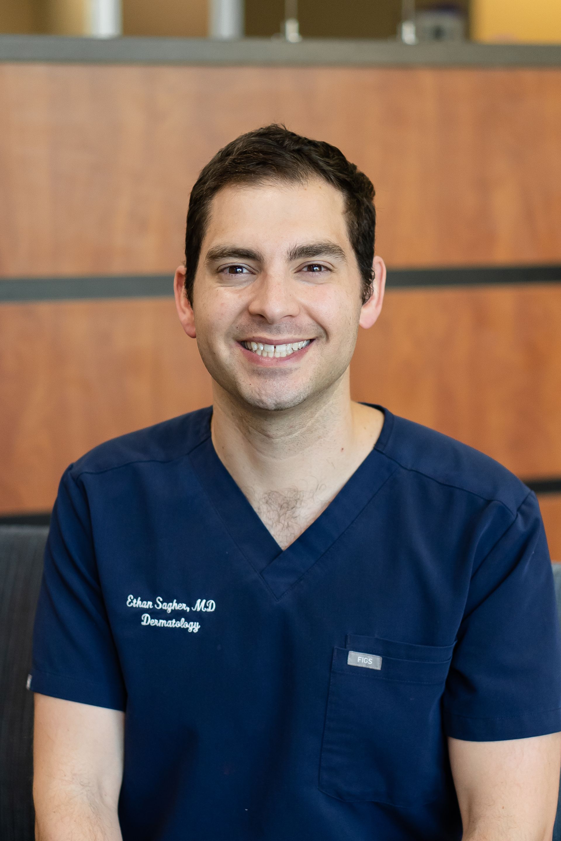 Man with brown hair smiles, wearing blue scrubs; office setting.