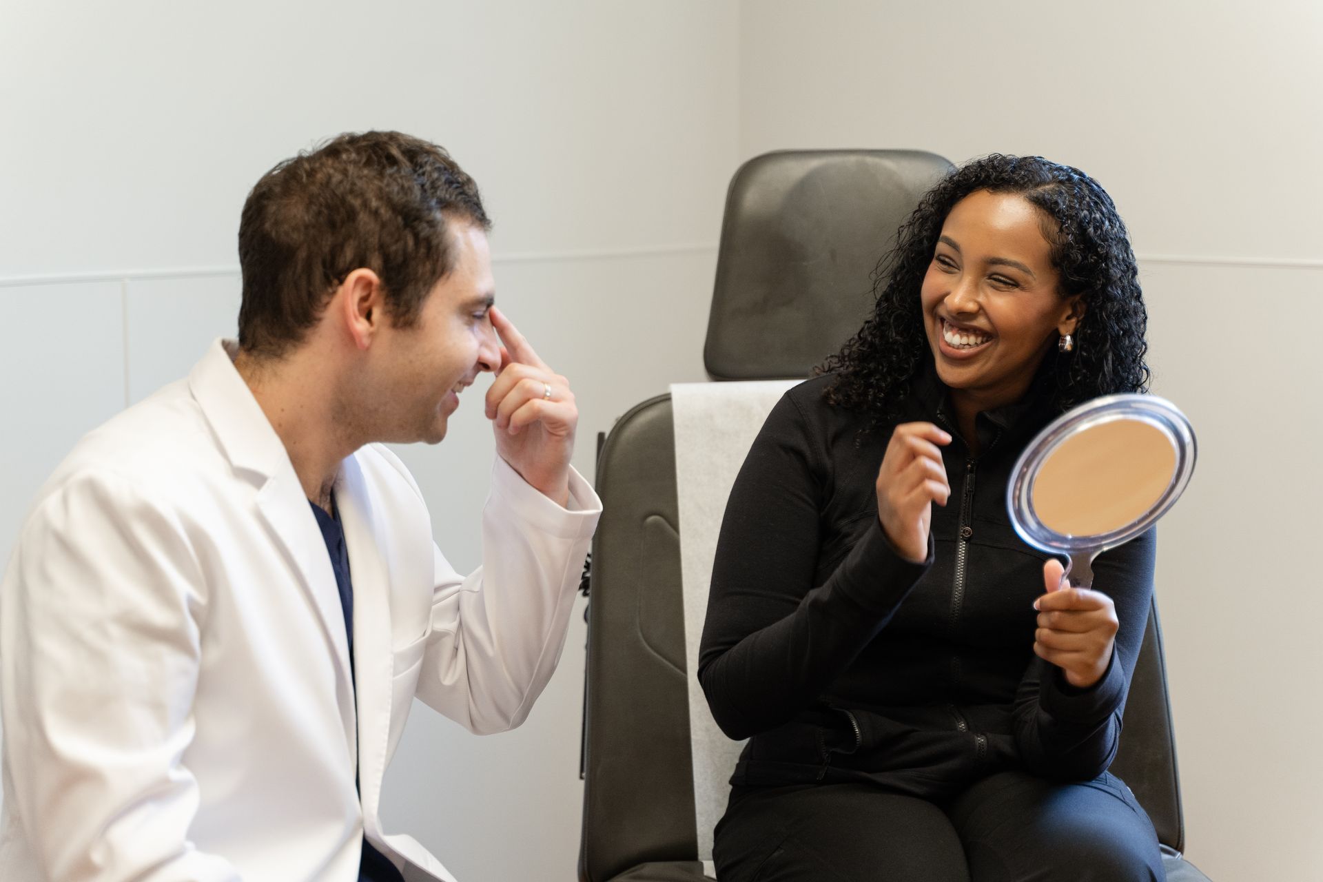 A doctor points to a woman's nose while she looks in a mirror, both smiling. Medical setting.