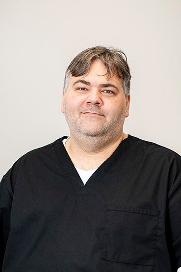 Man with dark hair, wearing black scrubs, smiling.