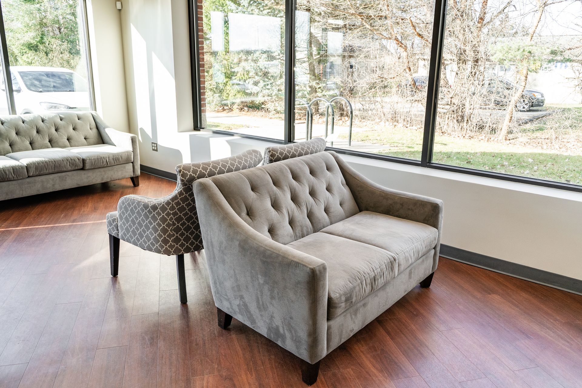 A waiting room with gray tufted couches and patterned chair, large windows, and hardwood floors.