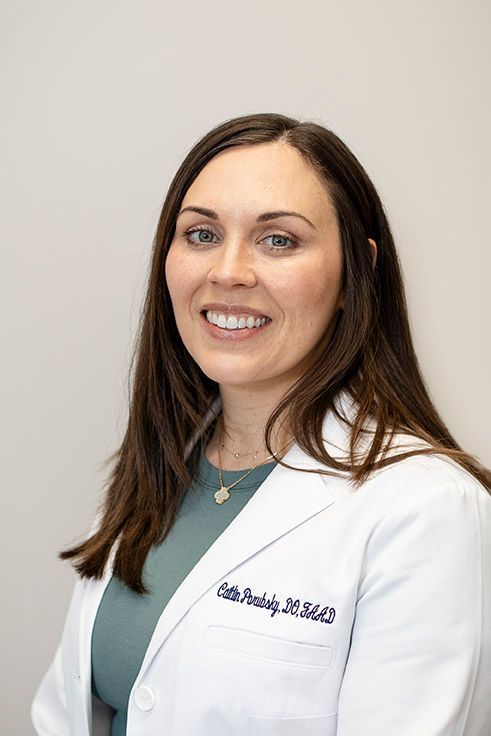 Woman in a white lab coat, smiles at the camera. She wears a green shirt, with a necklace, in a studio setting.