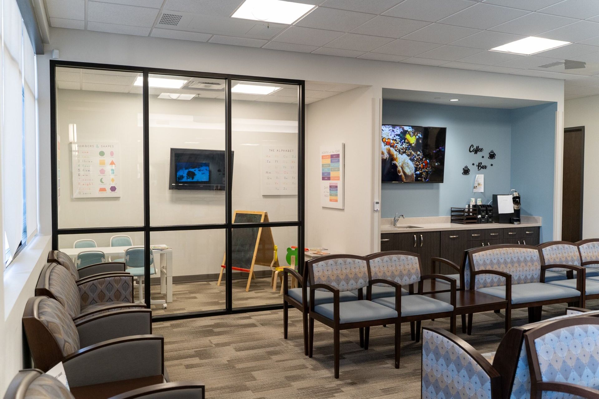 A waiting room with chairs, a play area behind glass, a TV, and coffee station.