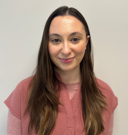 Woman with long brown hair smiling, wearing pink shirt, standing in front of a plain white background.