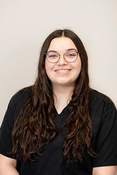 Woman with glasses smiles, wearing black scrubs, against a neutral wall.