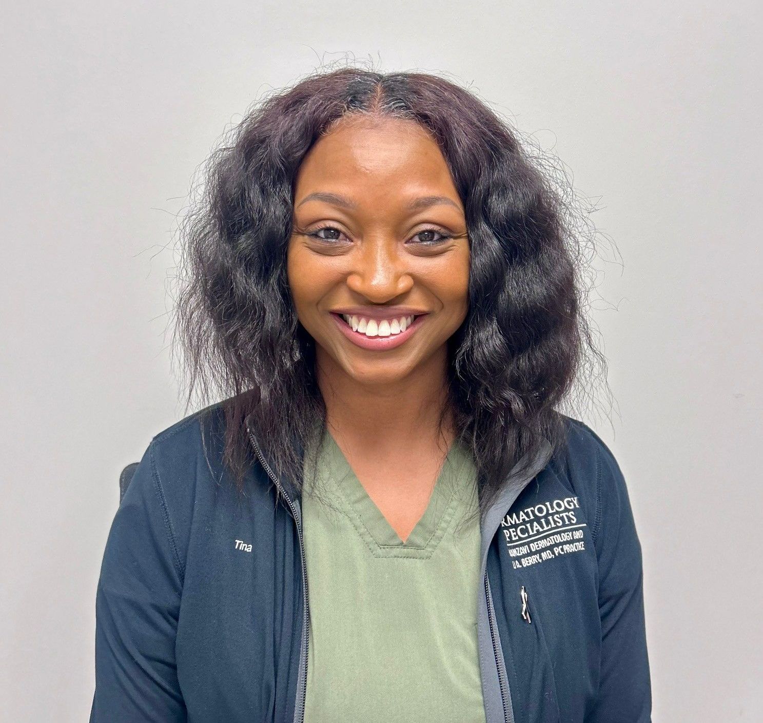 Smiling Black woman in scrubs; wears a black jacket, has dark curly hair, and stands in a medical setting.