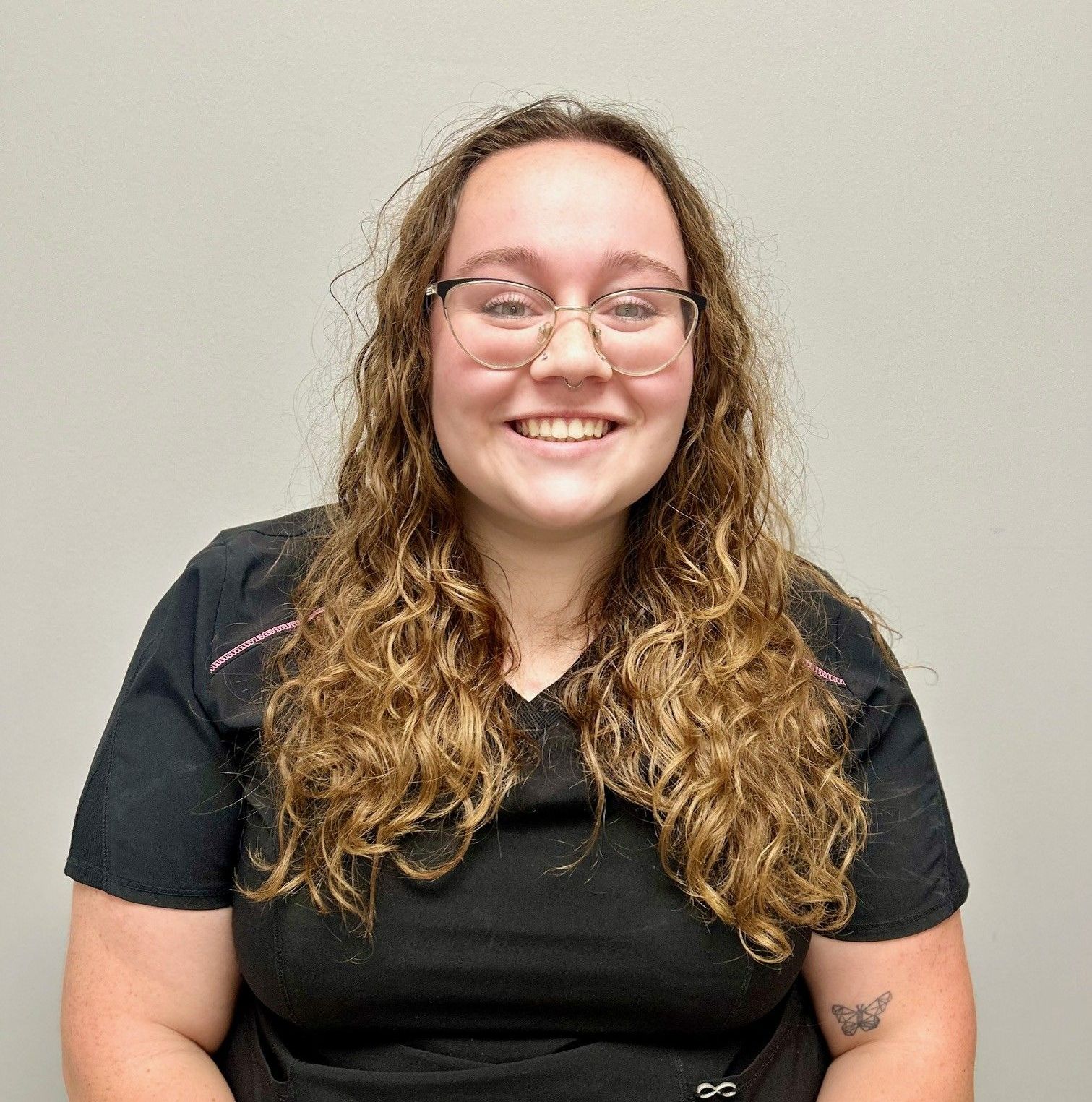 Woman with glasses, smiling, wearing black scrubs against a neutral background.