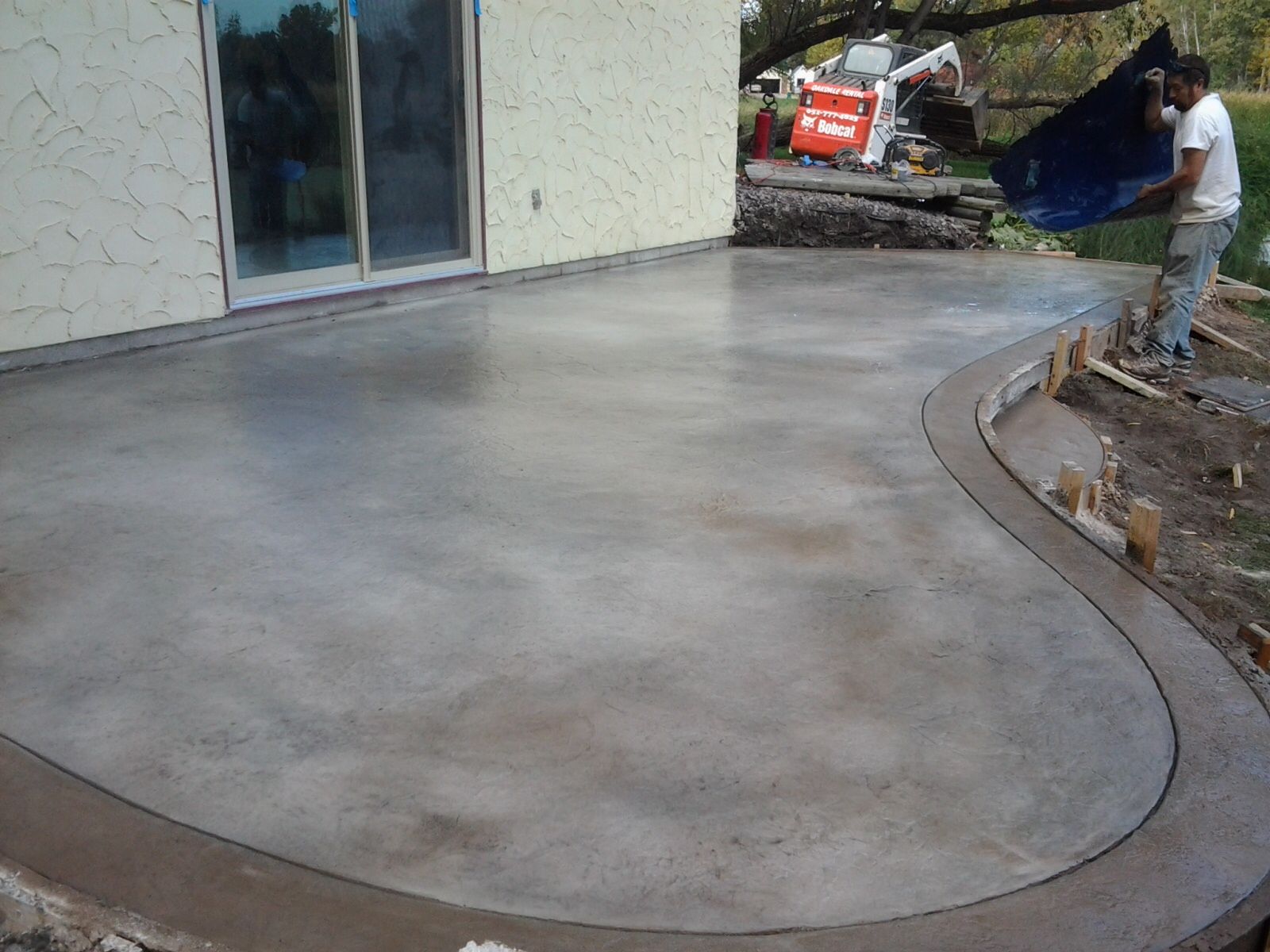 Concrete patio with curved edge; a person holds tarp near construction site.