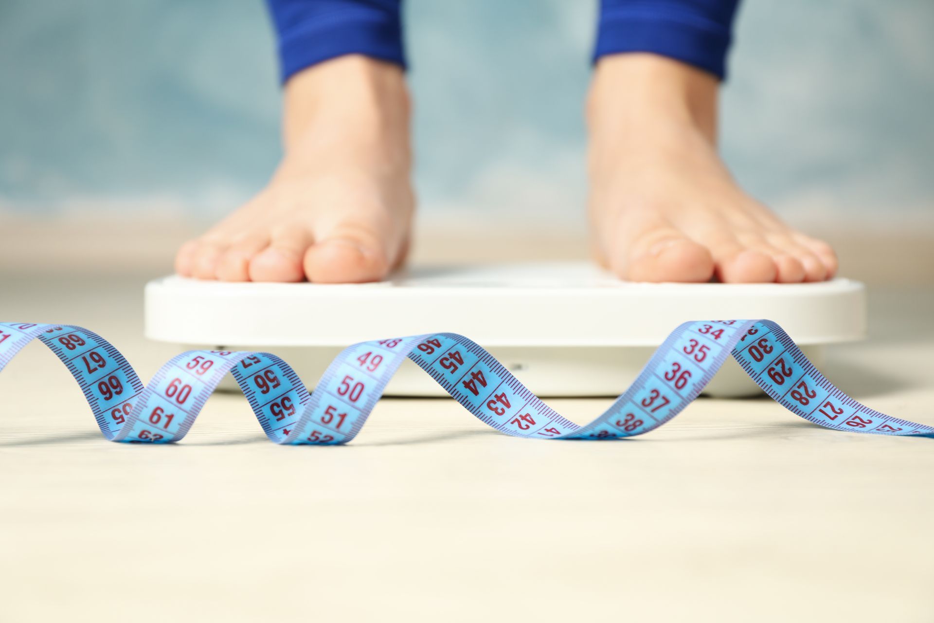 A person stands barefoot on a white digital scale, with a blue measuring tape coiled in the foreground.