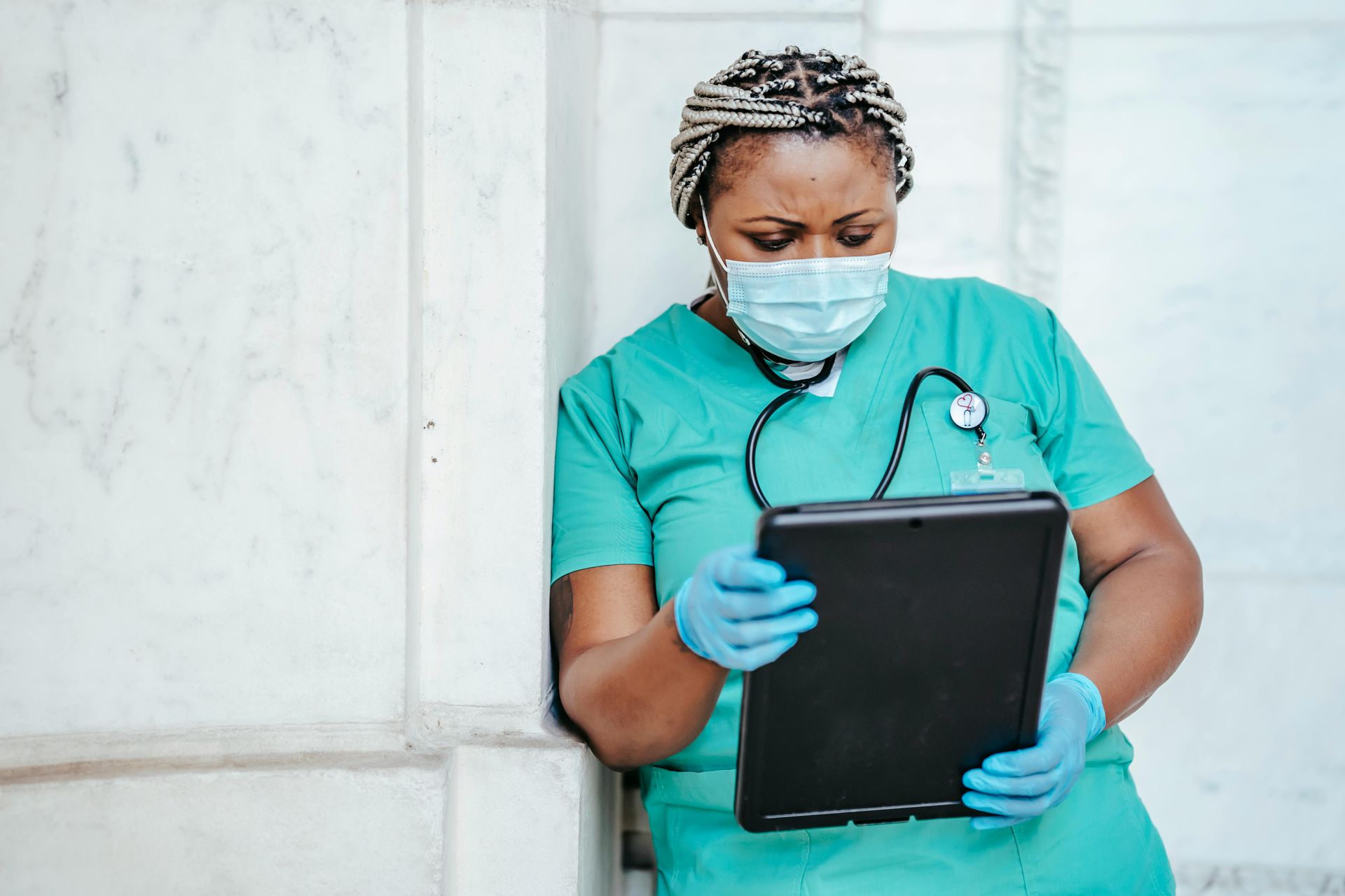 A medical worker in green scrubs, mask, and gloves, looks down at a clipboard, leaning against a white wall.